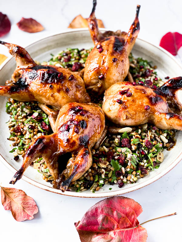 The Roasted Pomegranate Quail on a round white platter sitting on top of a Grain and Herb Salad. The platter is on a white marble benchtop with Autumn coloured leaves around the platter.