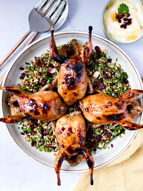 Looking down onto the round white platter with 4 Roasted Pomegranate Quail on top of a Grain and Herb Salad. Around the platter is a set of salad servers, a bowl of honey yoghurt and a mustard coloured napkin.