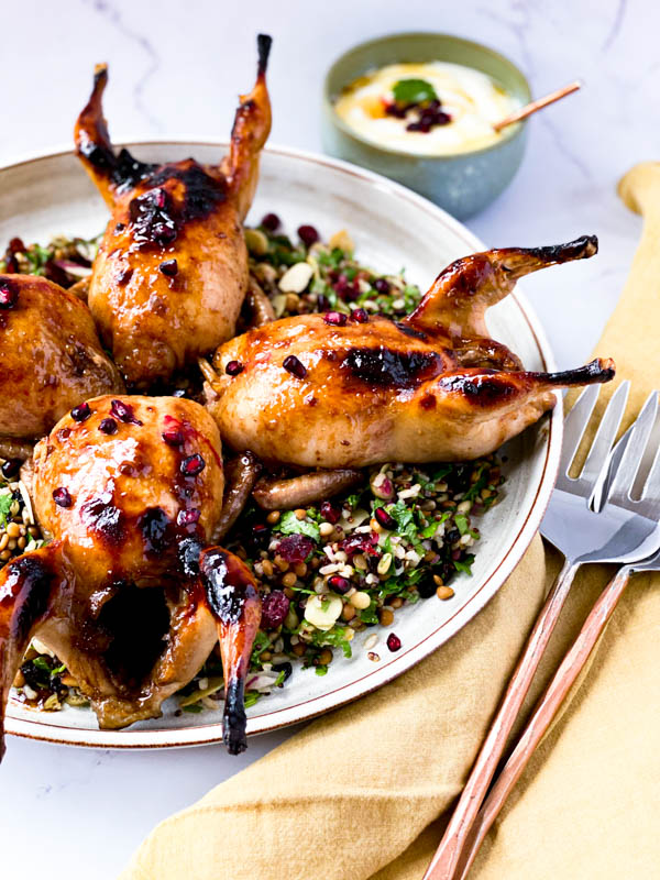 The white round platter with Roasted Pomegranate Quail sitting on top of a Grain and Herb Salad with salad servers and a napkin to the side and a small bowl of yoghurt dressing in the background.