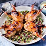 The 4 Roasted Pomegranate Quails sitting on a Grain and Herb Salad on a large white ceramic platter. It is surrounded by salad servers, grey napkins and a bowl of yoghurt dressing.