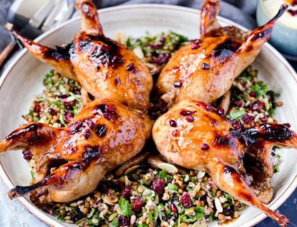 The 4 Roasted Pomegranate Quails sitting on a Grain and Herb Salad on a large white ceramic platter. It is surrounded by salad servers, grey napkins and a bowl of yoghurt dressing.