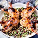 The Roasted Pomegranate Quail on a Grain and Herb Salad in a white round ceramic platter sitting on a dark marble table with grey napkins around it.