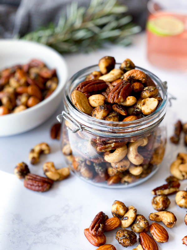 A jar of overflowing Sweet and Spicy Nuts with nuts scattered around it. A white bowl of nuts to the left and sprigs of thyme, a grey napkin and a glass of gin are in the background.
