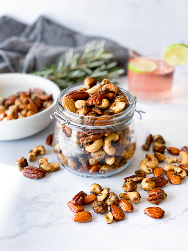 A jar of Sweet and Spicy Nuts in the centre without a lid on a white marble bench. Nuts are on the bench around it as well as in a white bowl to the left. Also in the background are a napkin, glass of gin and sprigs of rosemary.