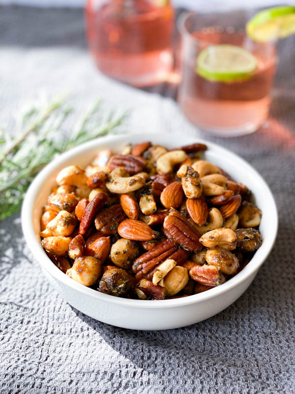 A white bowl of Sweet and Spicy Nuts in the centre on a grey tablecloth with sprigs of rosemary beside it and 2 glasses of gin in the background.