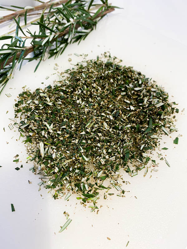 Finely chopped rosemary with a sprig in the background on a white bench.