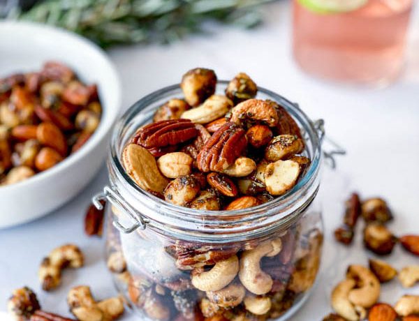 A jar and white bowl with Sweet and Spicy Nuts with nuts also on the white marble benchtop. Sprigs of rosemary and a glass of gin with a slice of lime are in the background.