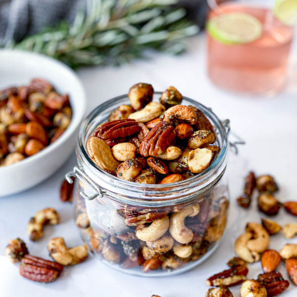 A jar and white bowl with Sweet and Spicy Nuts with nuts also on the white marble benchtop. Sprigs of rosemary and a glass of gin with a slice of lime are in the background.