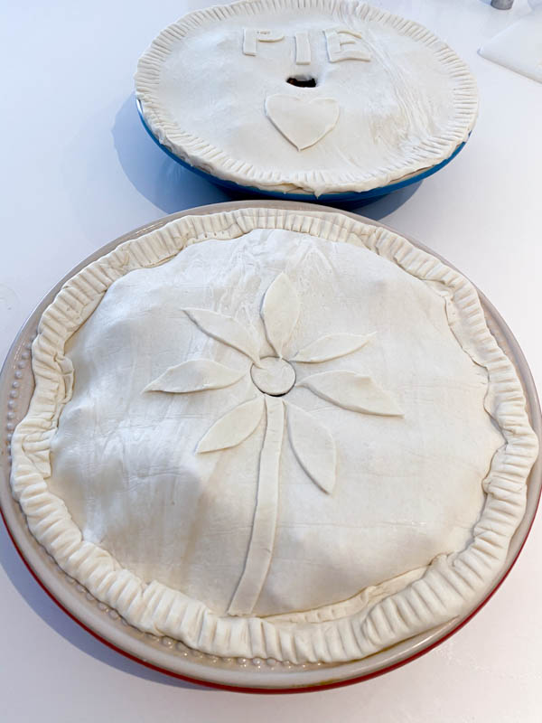 2 round pie dishes that are both uncooked and ready to go into the oven. One is decorated with a flower made of pastry and the other has the word 'pie' and a heart on it.