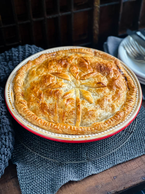 A cooked Beef and Vegetable family sized pie in a red ceramic pie dish, sitting on a wooden table on top of a wire resting rack.