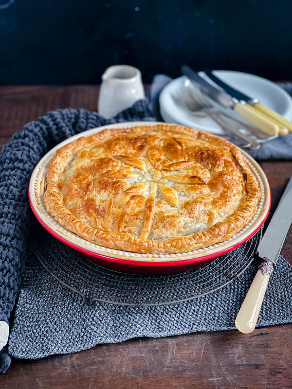 A family sized Beef and Vegetable Pie sitting on top of a resting rack on a dark wooden table with plates, cutlery and napkins in the photo.