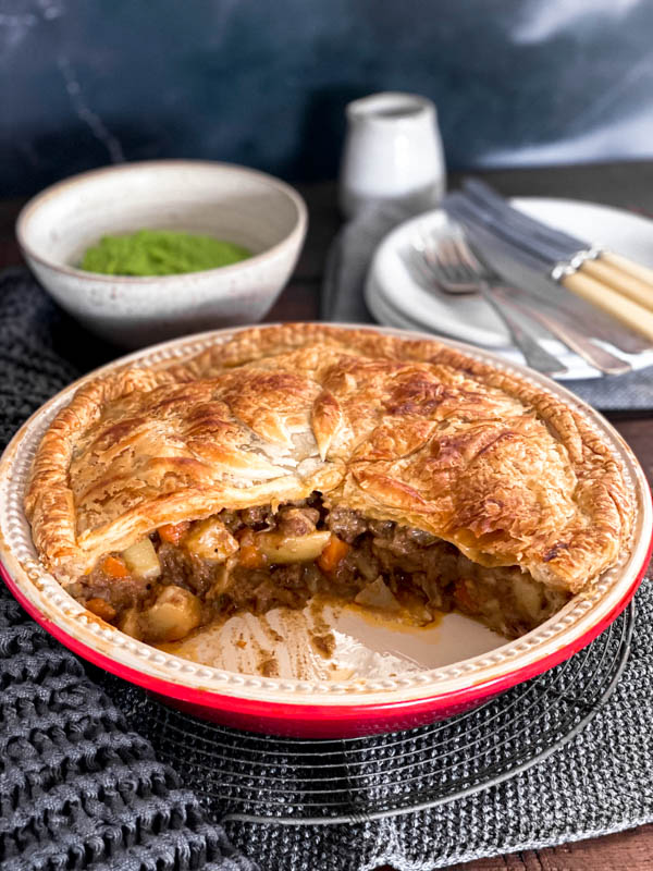 A family size Beef and Vegetable Pie that has had a wedge of pie removed so you can see the filling inside. Behind the pie are white plates, cutlery and a bowl of pea puree.