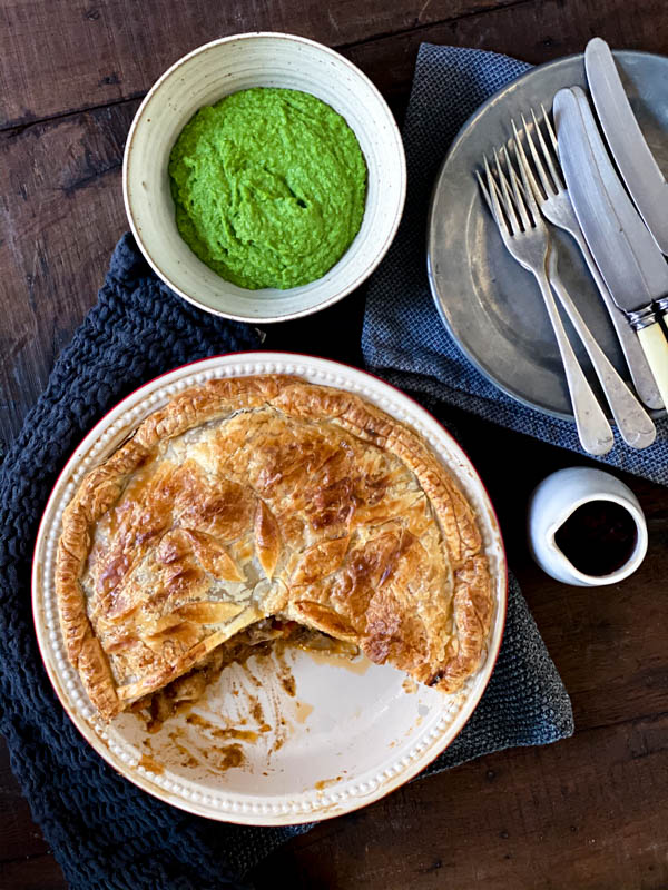 Looking down onto the Beef and Vegetable pie that has had a wedge taken from it, with a bowl of pea puree in the background. There is also a pewter plate with cutlery on top of it and a small jug of tomato sauce.