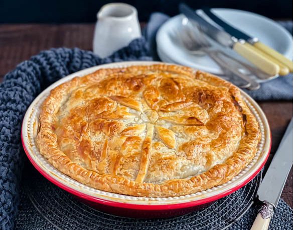 The family sized round Beef and Vegetable Pie on a dark wood table with white plates and cutlery in the background and a knife beside it.