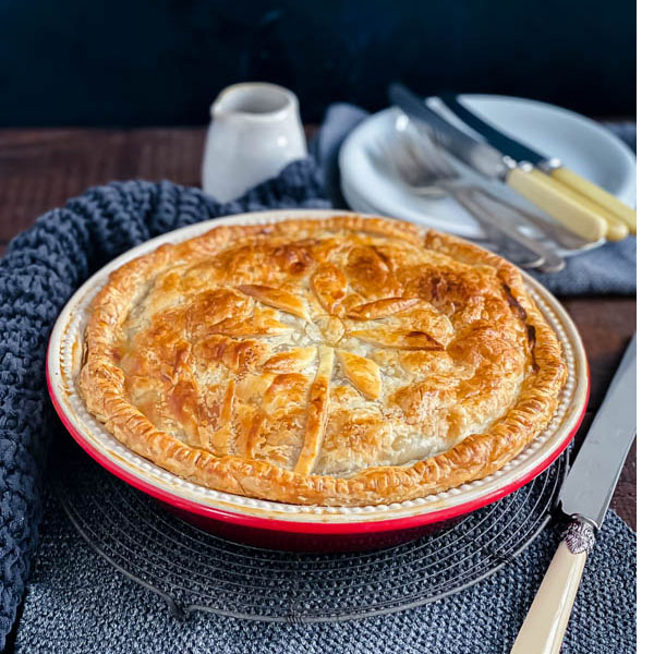 The family sized round Beef and Vegetable Pie on a dark wood table with white plates and cutlery in the background and a knife beside it.