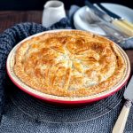 A round family sized Beef and Vegetable Pie on a round resting rack on top of a dark wood table with plates and cutlery in the background.