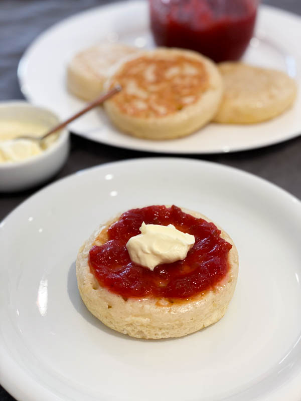 Close up of the Sourdough Crumpet with Rhubarb Compote and cream on top. In the background are more crumpets on a plate with the jar of compote and a small bowl of cream is in the middle.