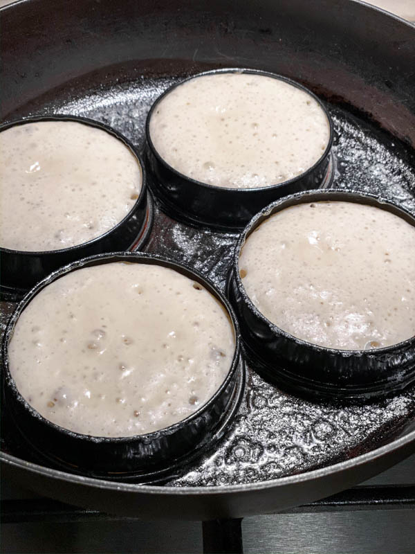 4 Sourdough Crumpets are being cooked. The batter is in crumpet rings with small holes appearing on the surface of the crumpets being cooked in a frying pan.