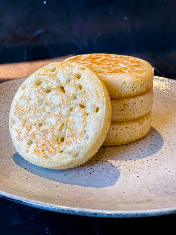 A stack of 3 sourdough crumpets with one leaning against them on a grey ceramic plate.