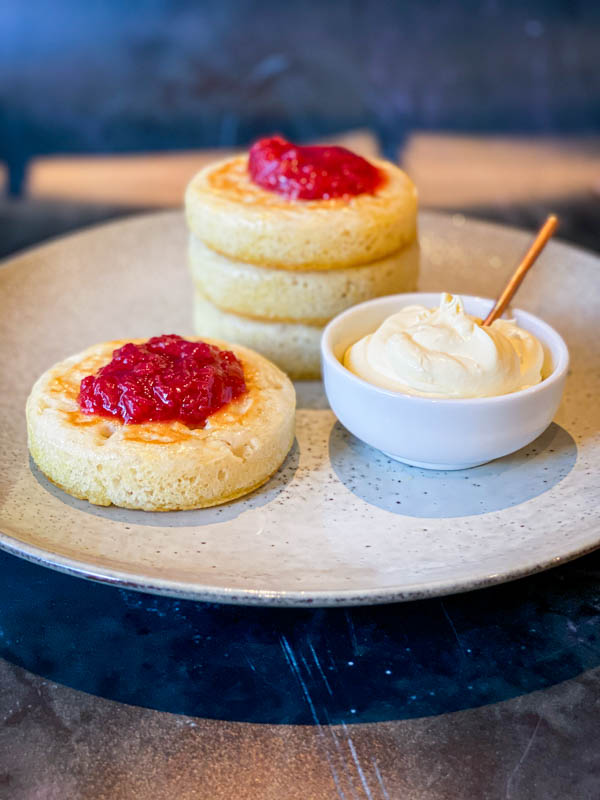 A stack of 3 sourdough crumpets on a grey plate with another crumpet in front of them. Both have a dollop of rhubarb compote on top and to the side, in a small bowl is cream fraiche with a small spoon sticking out of it.