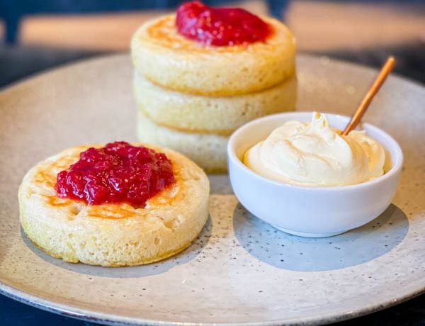 A grey ceramic plate with 3 stacked sourdough crumpets on it. In the front of them and still on the plate is another crumpet with a small bowl of creme fraiche. On top of the stack and on the single crumpet are dollops of rhubarb compote.