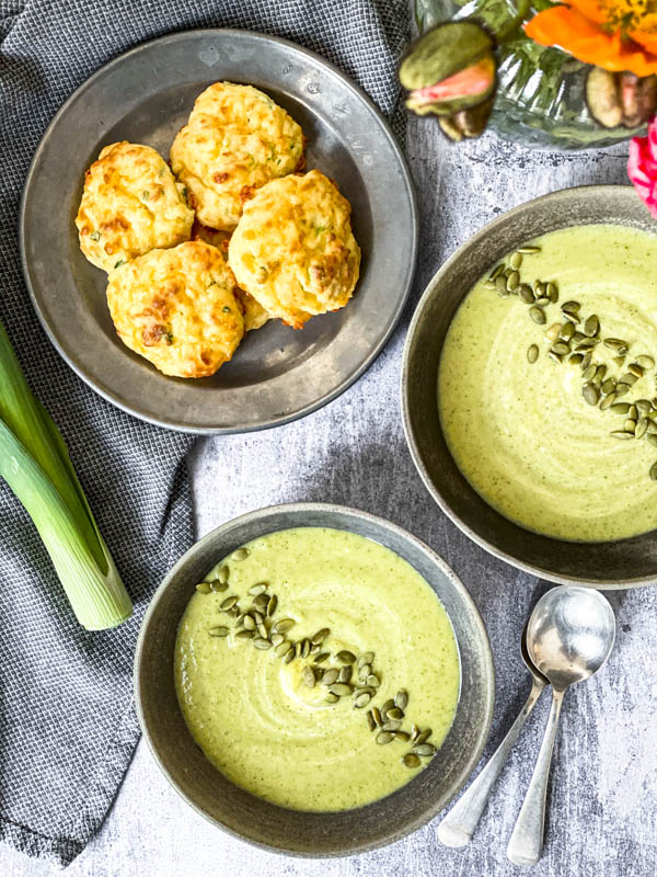 Looking down onto a table with 2 bowls of Broccoli Soup and a plate of Cheese Puffs. In the image are also 2 soup spoons, a napkin and just in the frame is a leek stalk and a vase of poppies.