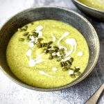 A grey ceramic bowl with Broccoli Soup in it. It is garnished with a line of pumpkin seeds down the middle and a swirl of cream.