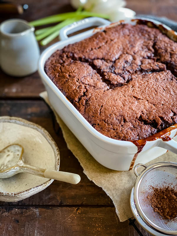 Chocolate Self Saucing Pudding in a white square dish on a wooden table with a ceramic bowl and spoon next to it. A small sieve with cocoa is in the front and a small ceramic cream jug is in the background.