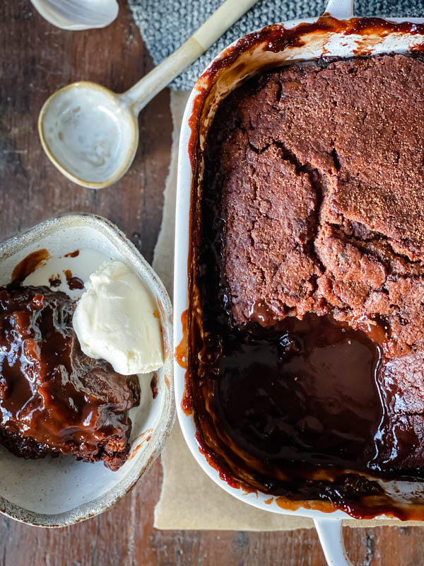 Looking down onto a white oven dish with Chocolate Self Saucing Pudding inside. Next to it is a ceramic bowl filled with a serving of pudding and a scoop of vanilla ice-cream.