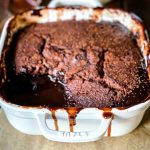 Chocolate Self Saucing Pudding in a square, white ovenproof dish with a spoonful taken out to reveal the chocolate sauce in the bottom of it. It is sitting on top of brown baking paper on a wire resting rack.