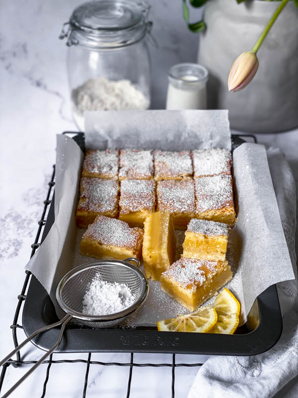 A tray of Gooey Yuzu Slice cut into squares with a small sieve of icing sugar resting on the tray. In the background are a jar of flour and a small beaker of milk.