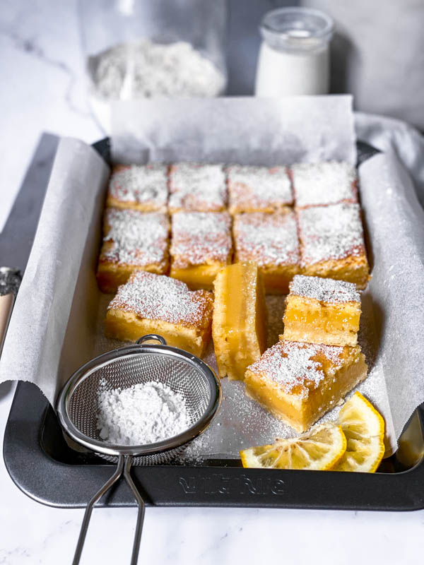 A tray of Gooey Yuzu Slice that is cut into squares and dusted with icing sugar. In the foreground is a small sieve with icing sugar and 2 yuzu slices cut in half.