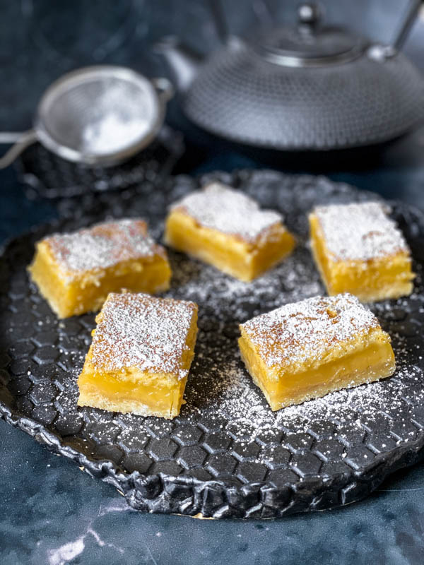 5 squares of Gooey Yuzu Slice dusted with icing sugar sitting on a textured black plate. In the background is a black japanese teapot and a small sieve.