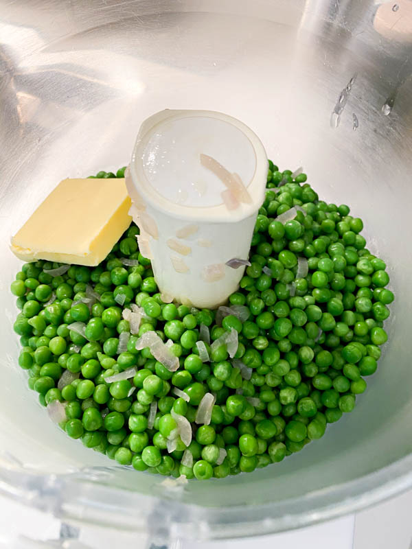 The cooked baby peas and onions are in the bowl of a food processor with a piece of butter, ready to be processed.