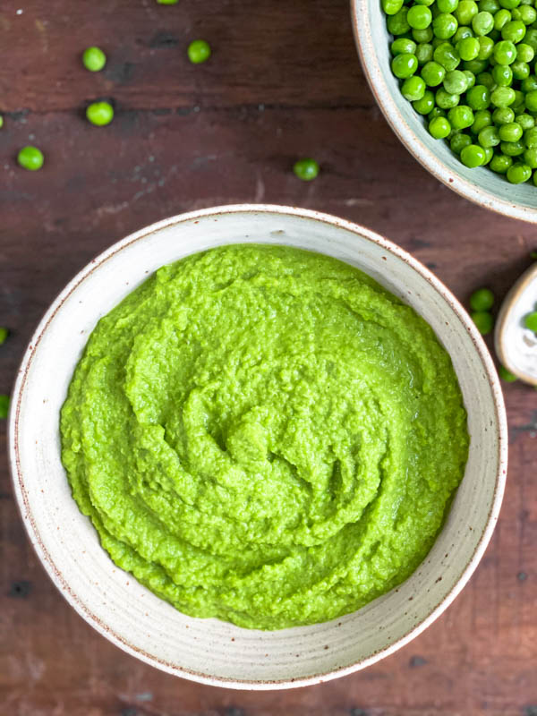 Looking down into a ceramic bowl of green Pea Purée on a wooden table with a few peas scattered around it. There is also a bowl of green baby peas behind it in the top corner.