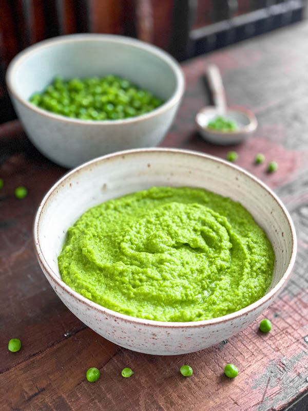A white speckled ceramic bowl full of vibrant green Pea Purée. There are a few baby peas scattered around the bowl on a wooden table and another bowl of whole baby green peas and a spoon in the background.