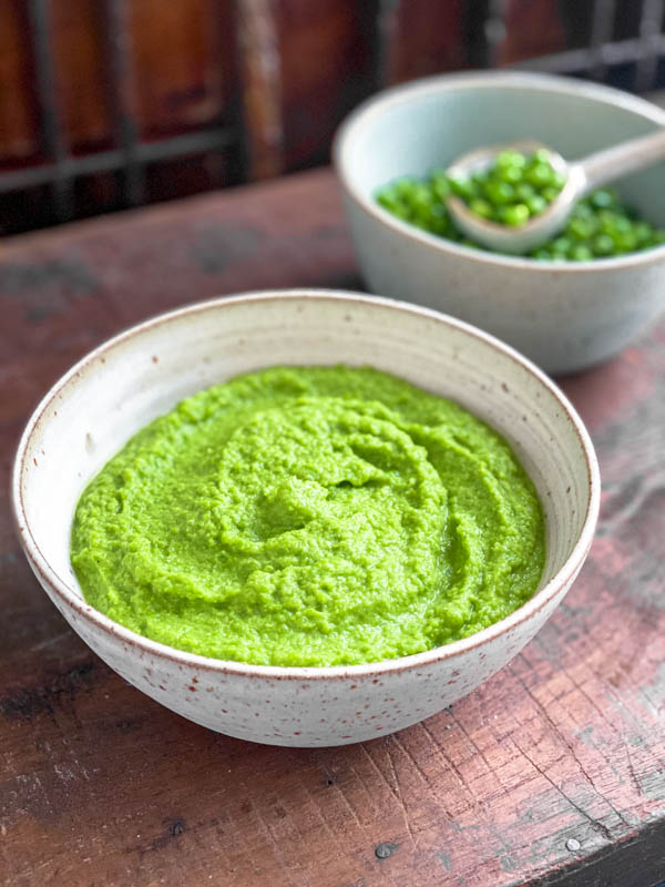 A bowl of green Pea Purée with another bowl of whole peas in the background. They are on a dark wood table.