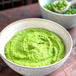 A bowl of Pea Purée in the front, with a bowl of whole green peas behind it with a spoon in the bowl.