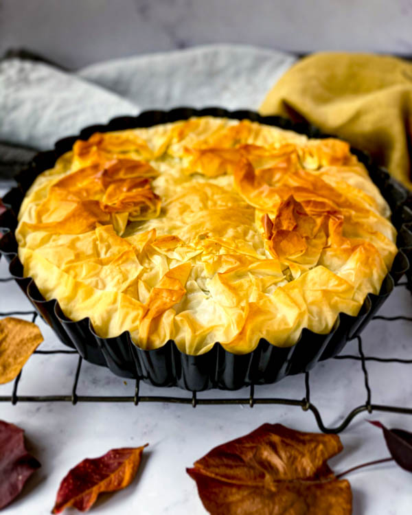 A Spanakopita Pie baked in a round Tart Tin sitting on a black resting rack with Autumn leaves around it and napkins in the background.