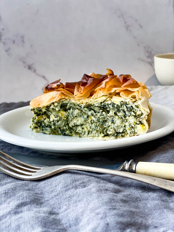 A slice of Spanakopita Pie on a white plate with a knife and fork in front of the plate. The plate is on a grey blue linen tablecloth.