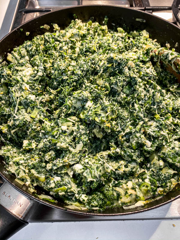 The Spanakopita Pie filling of spinach, cheeses, onions, leeks and herbs are all mixed together in a frying pan ready for filling the pie.