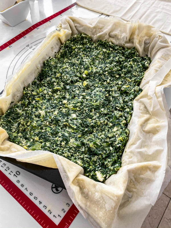 The filo pastry sheets are buttered and laid out in a rectangle baking dish with the Spanakopita Pie filling inside, ready for the top layers of filo to be added.