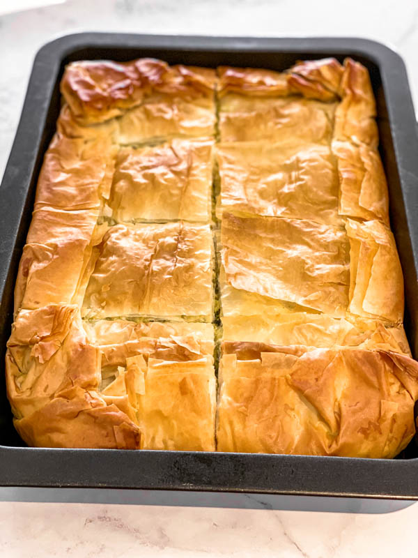 The Spanakopita Pie is golden and ready. It is in a rectangle baking dish and is cut into 8 pieces but still in the dish.