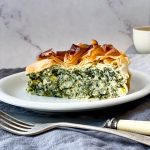 A slice of Spanakopita Pie sitting on a white small plate with a knife and fork in the foreground and draped linen napkins in the background.