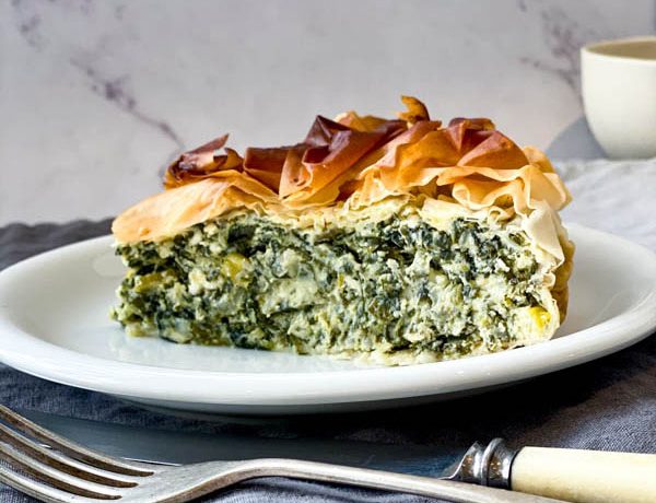 A slice of Spanakopita Pie sitting on a white small plate with a knife and fork in the foreground and draped linen napkins in the background.