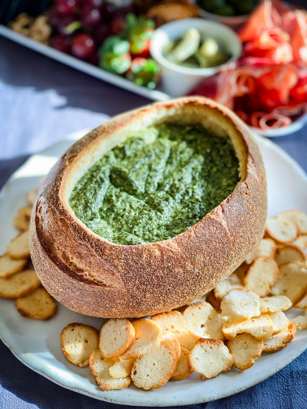 The Spinach Cobb Loaf Dip is on a platter, with bagel crisps scattered around it. In the background is a cheese and meat platter.