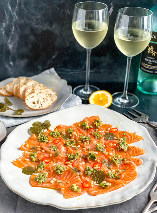 A close up of the platter of thinly sliced sashimi salmon topped with dollops of the chunky tartare mix. In the background are 2 glasses of wine, baguette slices and a lemon half, all on a black marble bench.