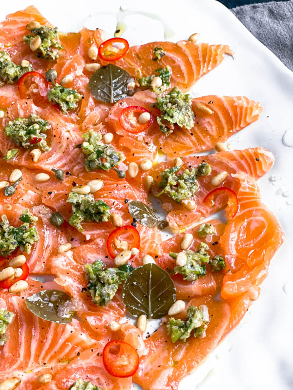 A close up of the thinly sliced salmon sashimi with dollops of tartare on top of the salmon pieces, along with slices of red chilli, pinenuts and caper berries and leaves.