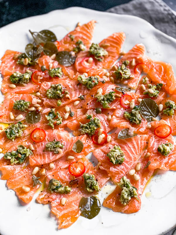 The platter of thinly sliced salmon sashimi pieces with the dollops of tartare on top. It also has sliced red chillies, pinenuts, capers and caper leaves scattered over the salmon. The whole plate is drizzled with olive oil.