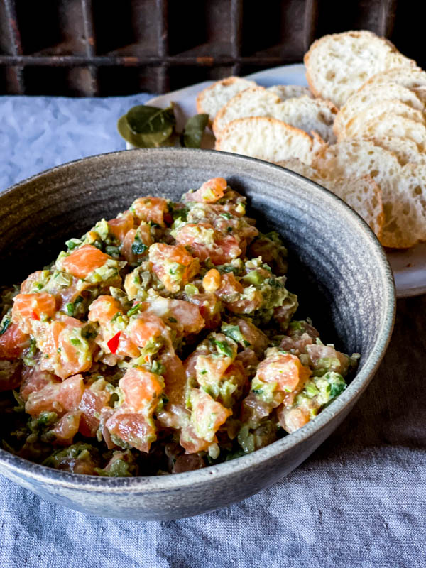 A ceramic bowl of Salmon Tartare with some thinly sliced baguette rounds behind it on a plate.